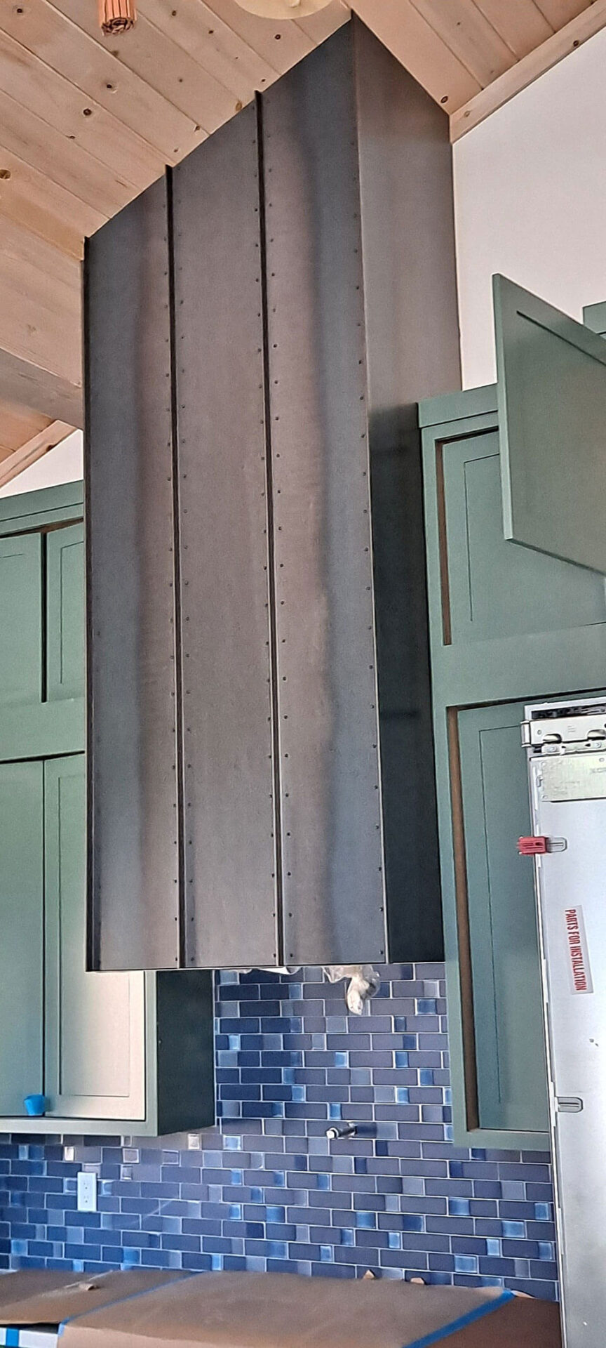 A large dark metal range hood extends from a wood-paneled ceiling above blue tiled backsplash and green kitchen cabinets.