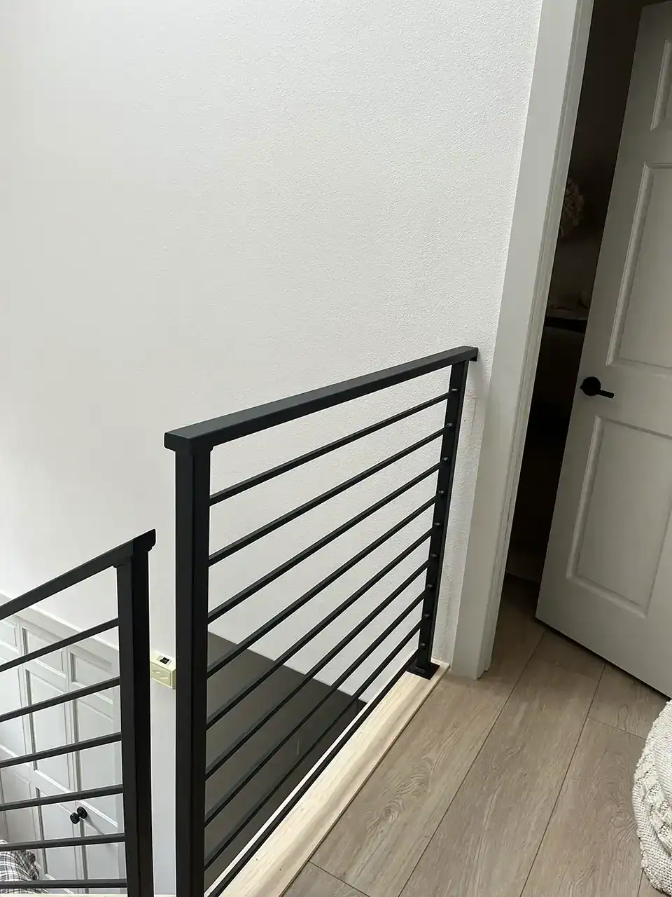 black metal stair railing next to a beige wall and wooden floor, with an open white door leading to another room in the background.