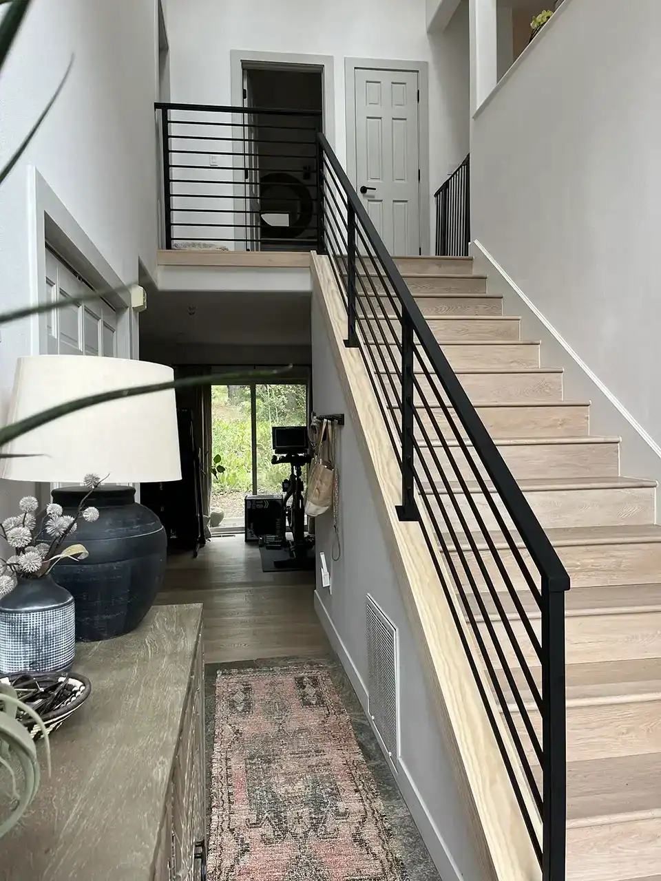 a modern staircase with black metal railings leads to an upper floor; a hallway and doorways are visible, with a console table and decor in the foreground.