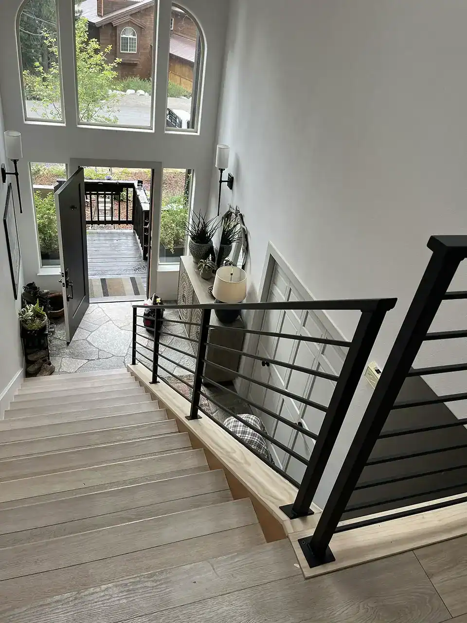 view from the top of a staircase overlooking a modern entryway with an open front door, large windows, and indoor plants.