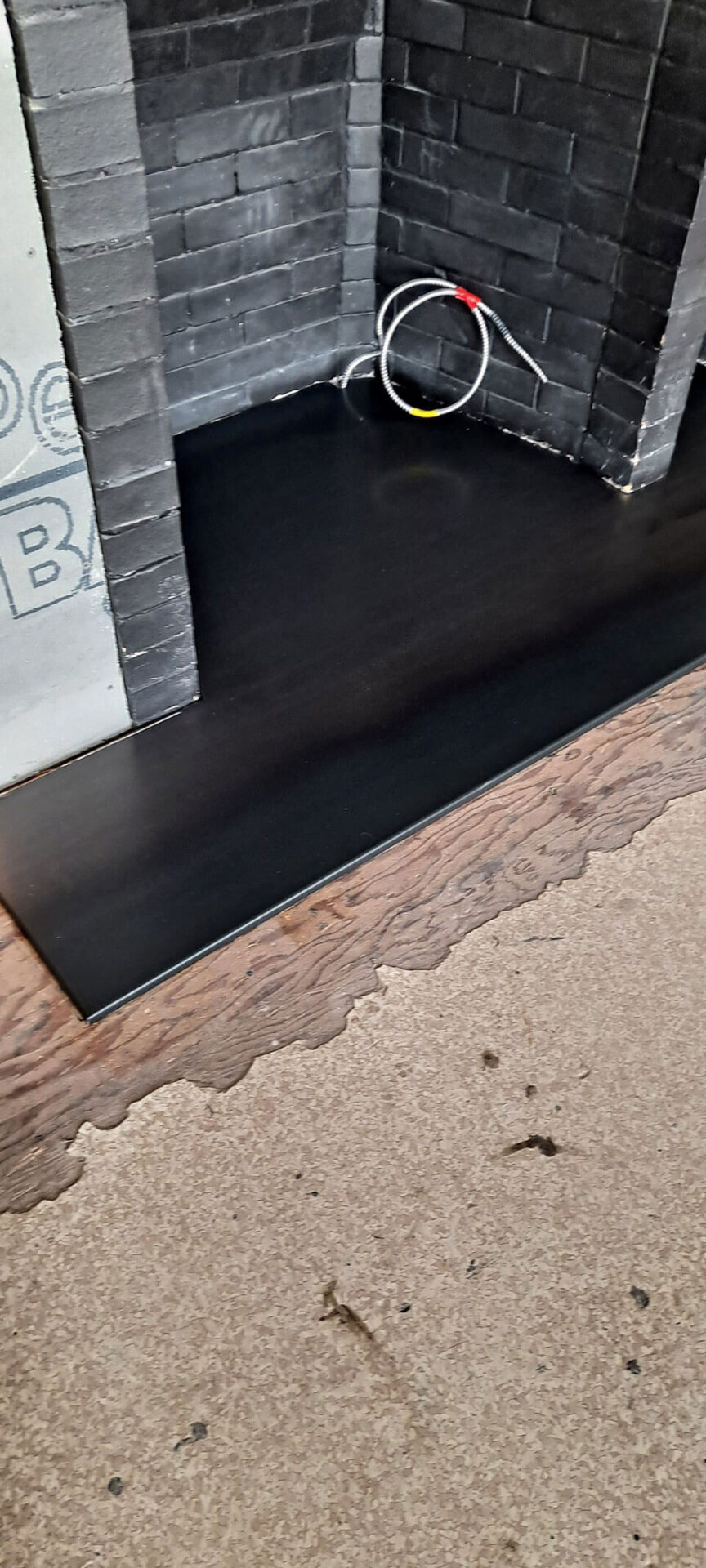 A black panel covers part of a floor near a fireplace with exposed wiring; the floor transitions from wood to a worn carpet.