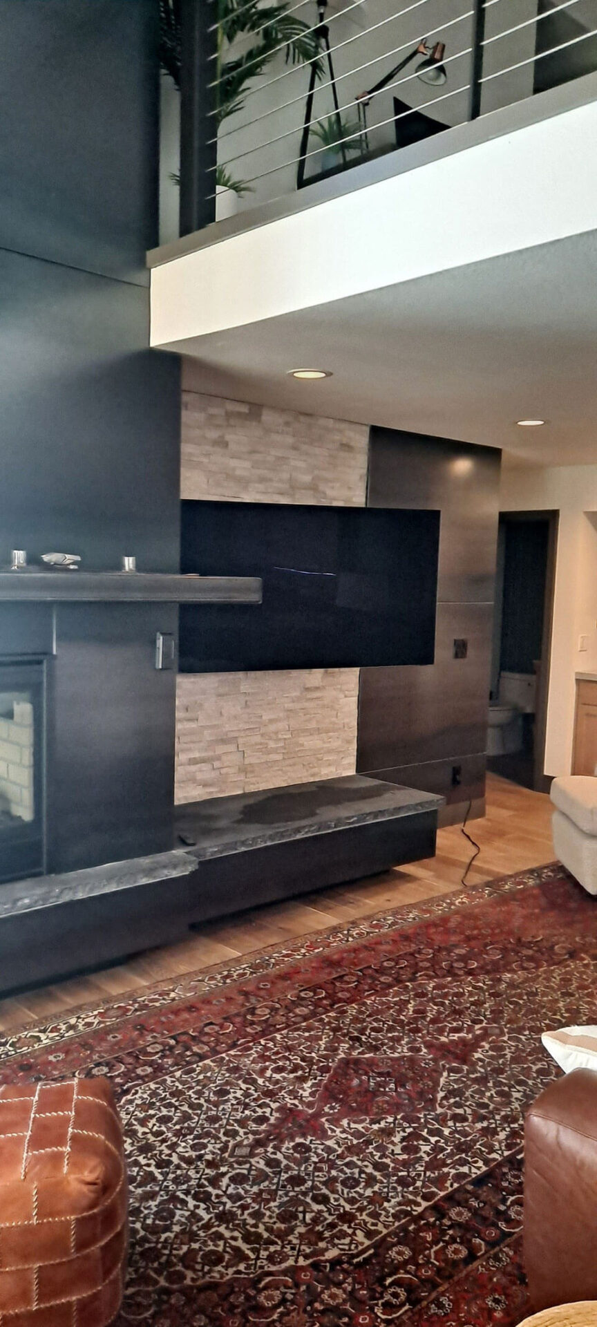 Modern living room with a wall-mounted TV, stone accent wall, dark wood paneling, fireplace, and a patterned rug; partial view of an upstairs railing and furniture.
