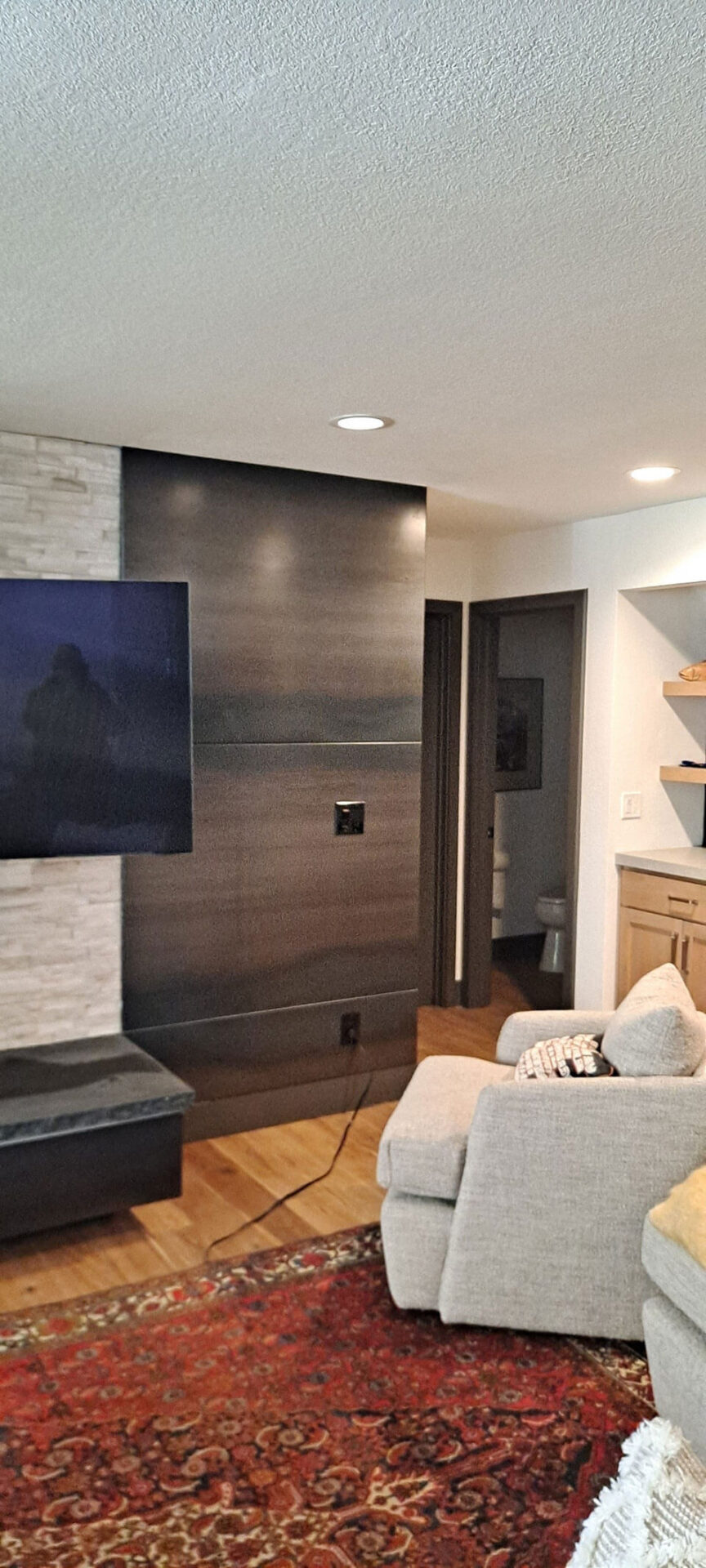 A living room with a mounted TV on a dark accent wall, a light-colored armchair, a red patterned rug, and an open door revealing a bathroom.
