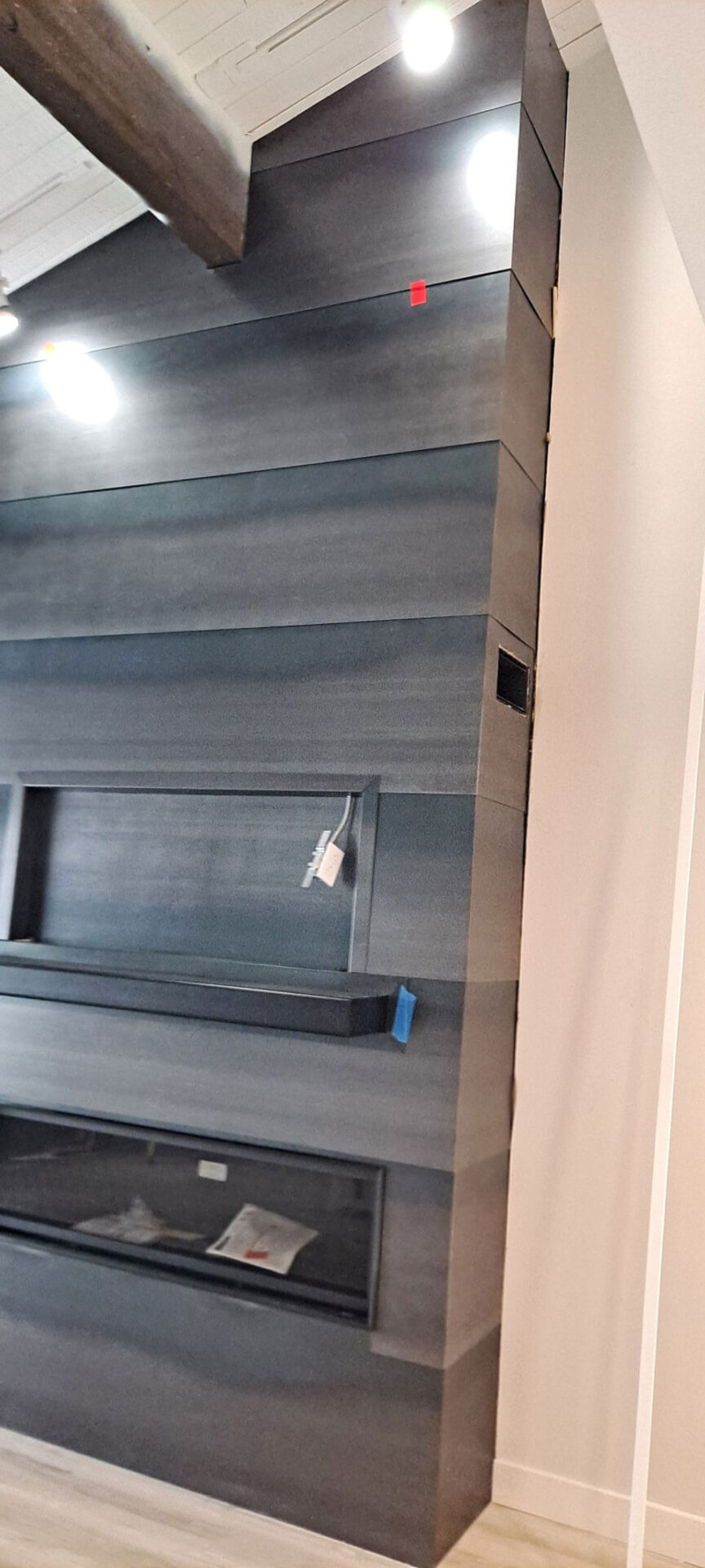 A modern, dark wood-paneled wall with integrated shelving, a built-in fireplace, ceiling lights, and some visible construction tags.
