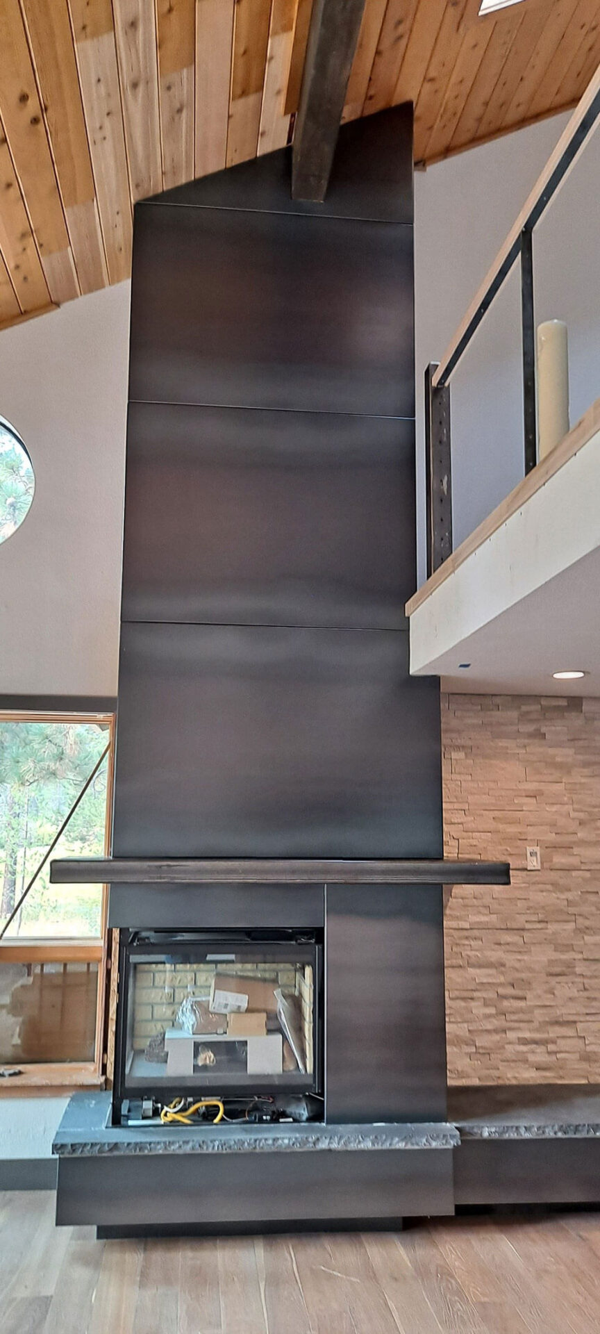 A modern double-sided fireplace with a tall, matte black metal surround, set in a room with wood ceilings and stone accent walls.