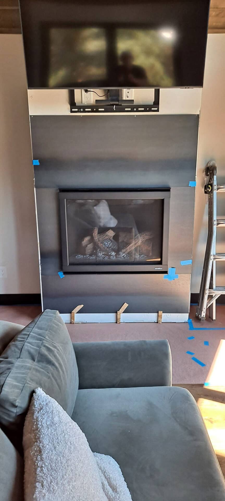 A living room with a fireplace under construction, blue painter’s tape on the wall, a TV mounted above, a ladder to the right, and part of a couch with a white pillow in the foreground.
