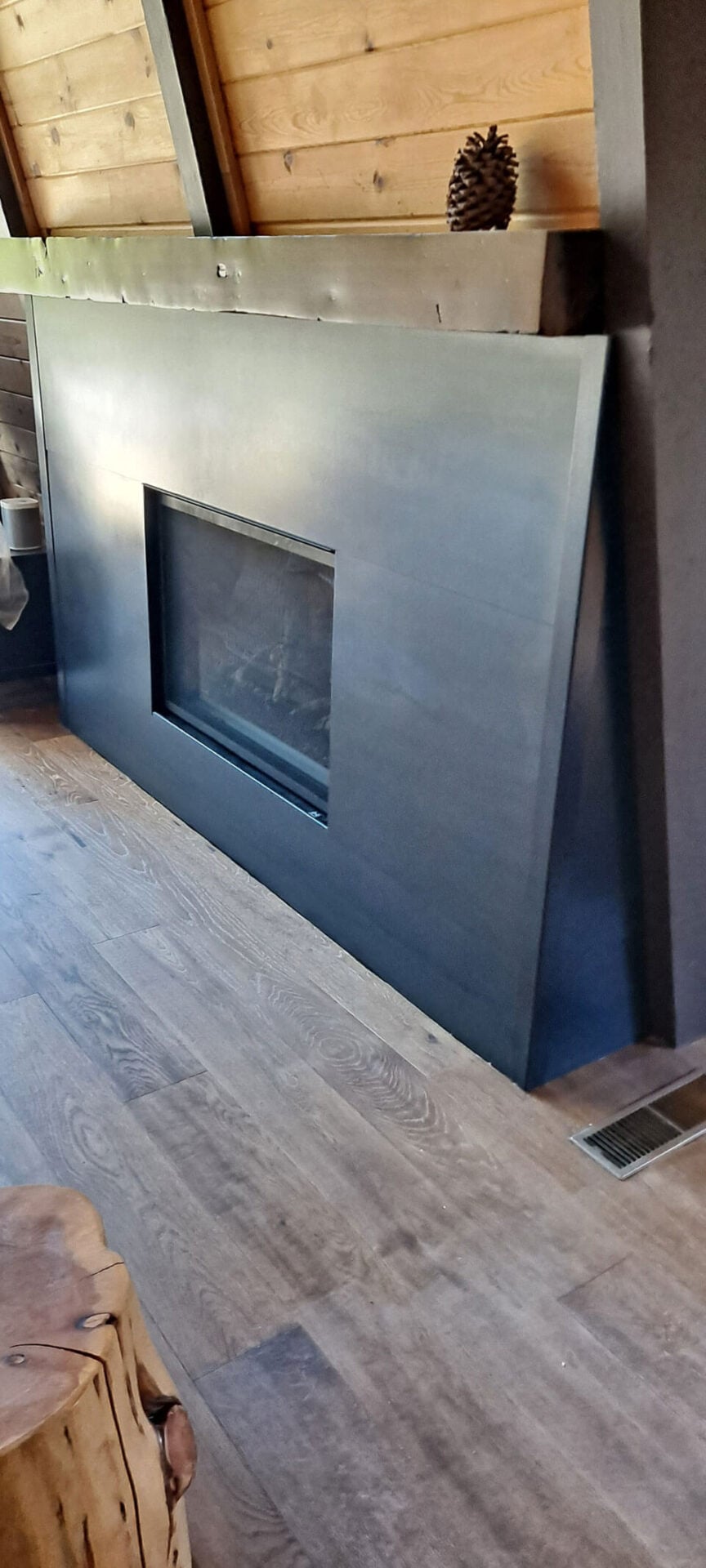 Modern fireplace with a matte black, angled metal surround set against wood-paneled walls and flooring; a pine cone sits on top.