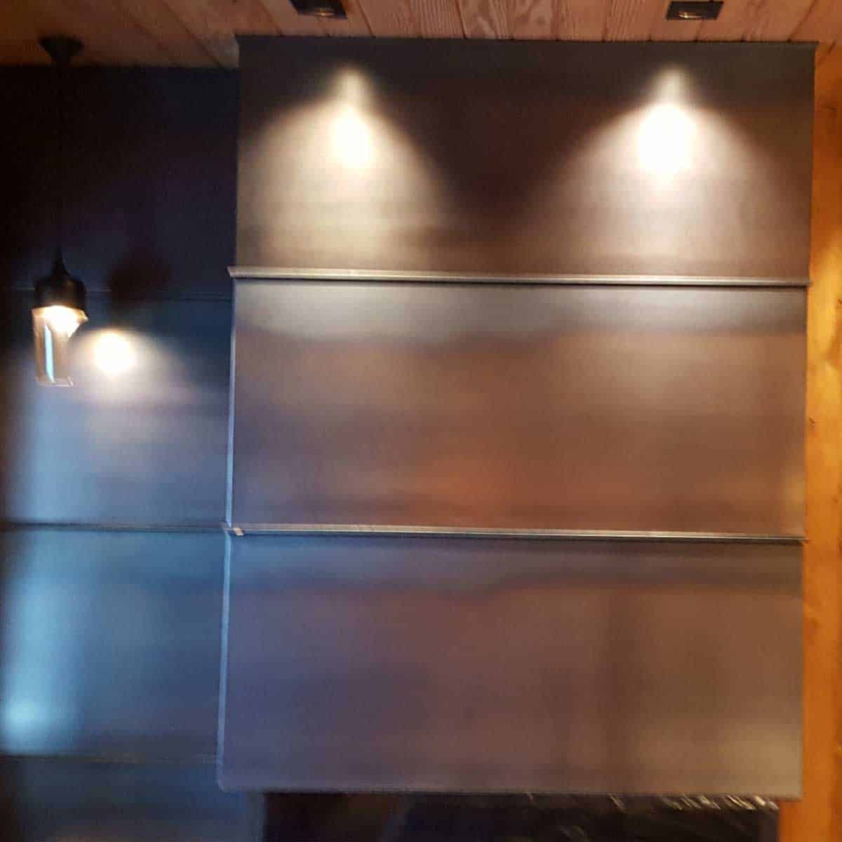 A wall with three large horizontal metal panels, illuminated by three ceiling spotlights and a hanging pendant light on the left side.