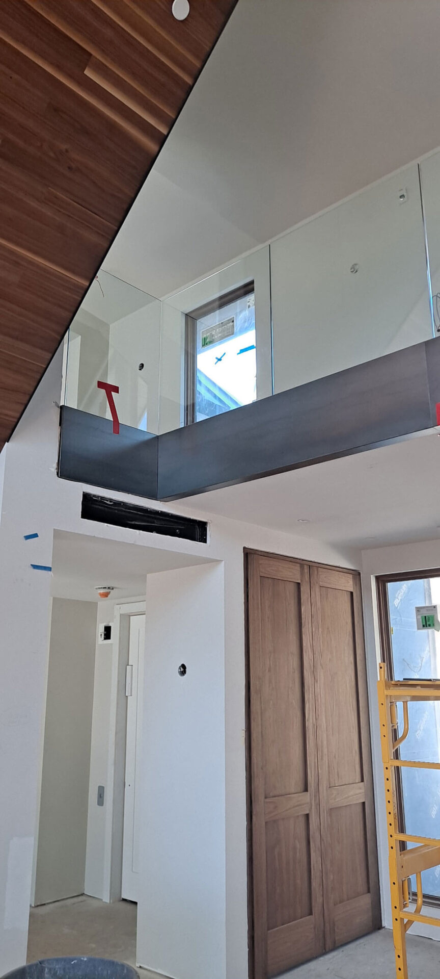 Interior view of a modern building under construction, featuring glass railings, wooden ceiling panels, a wooden sliding door, and visible construction materials.