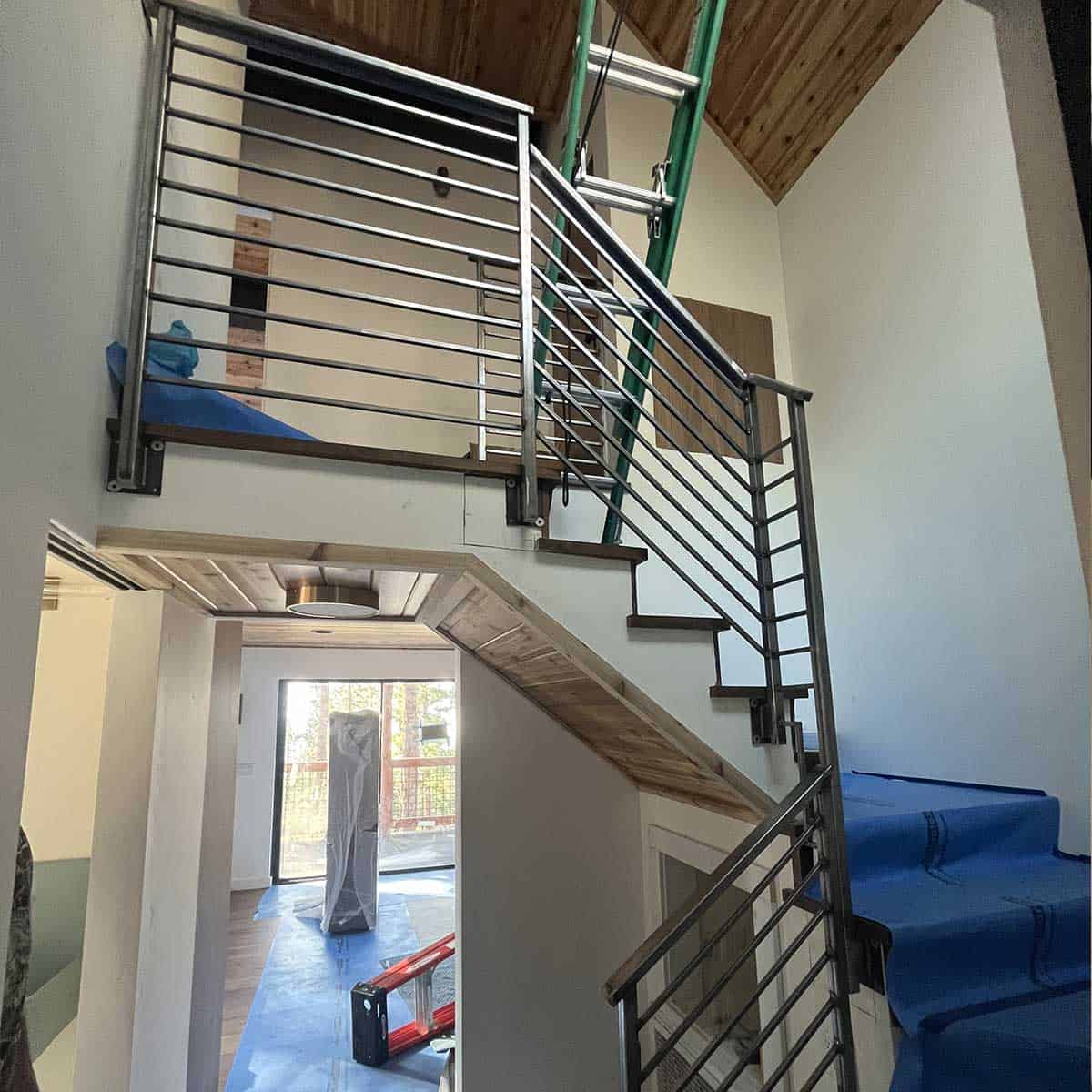 A modern indoor staircase with metal railings and blue protective coverings on the steps, leading to an upper level under a wooden ceiling.