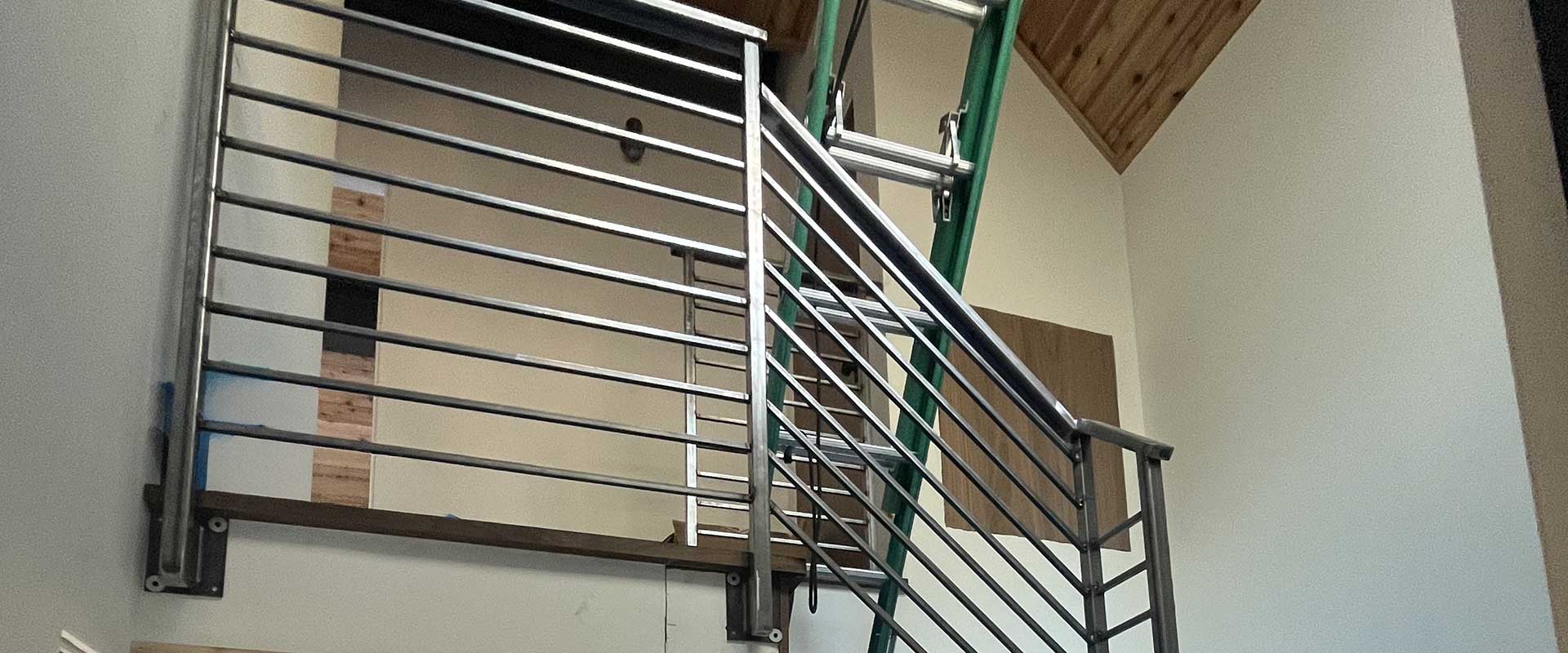 Metal stair railing with horizontal bars and a green ladder positioned behind it, inside a room with white walls and a wooden ceiling.
