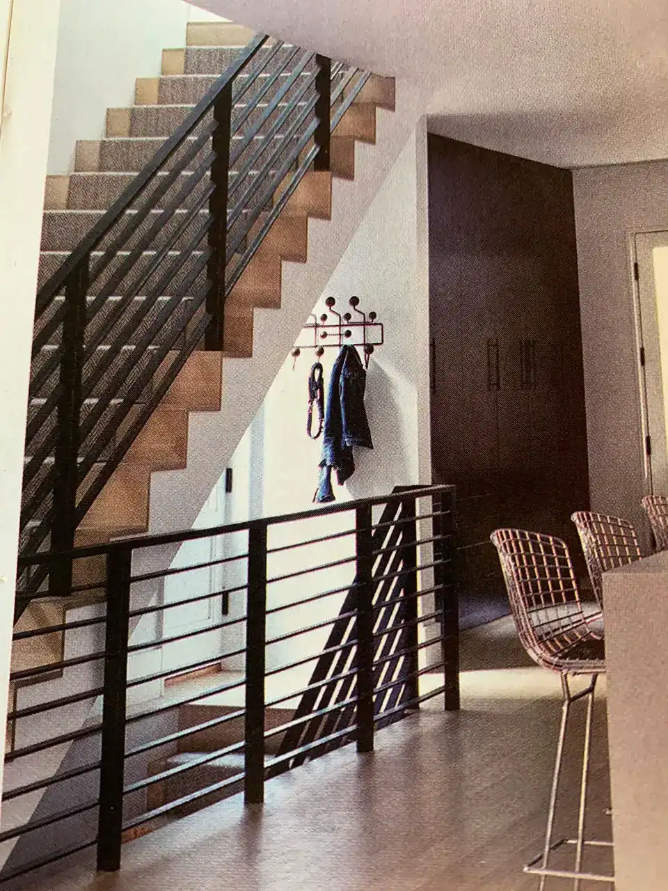 modern interior with a wooden staircase and black metal railings, a coat rack with jackets, tall chairs at a counter, and natural light from a nearby window.