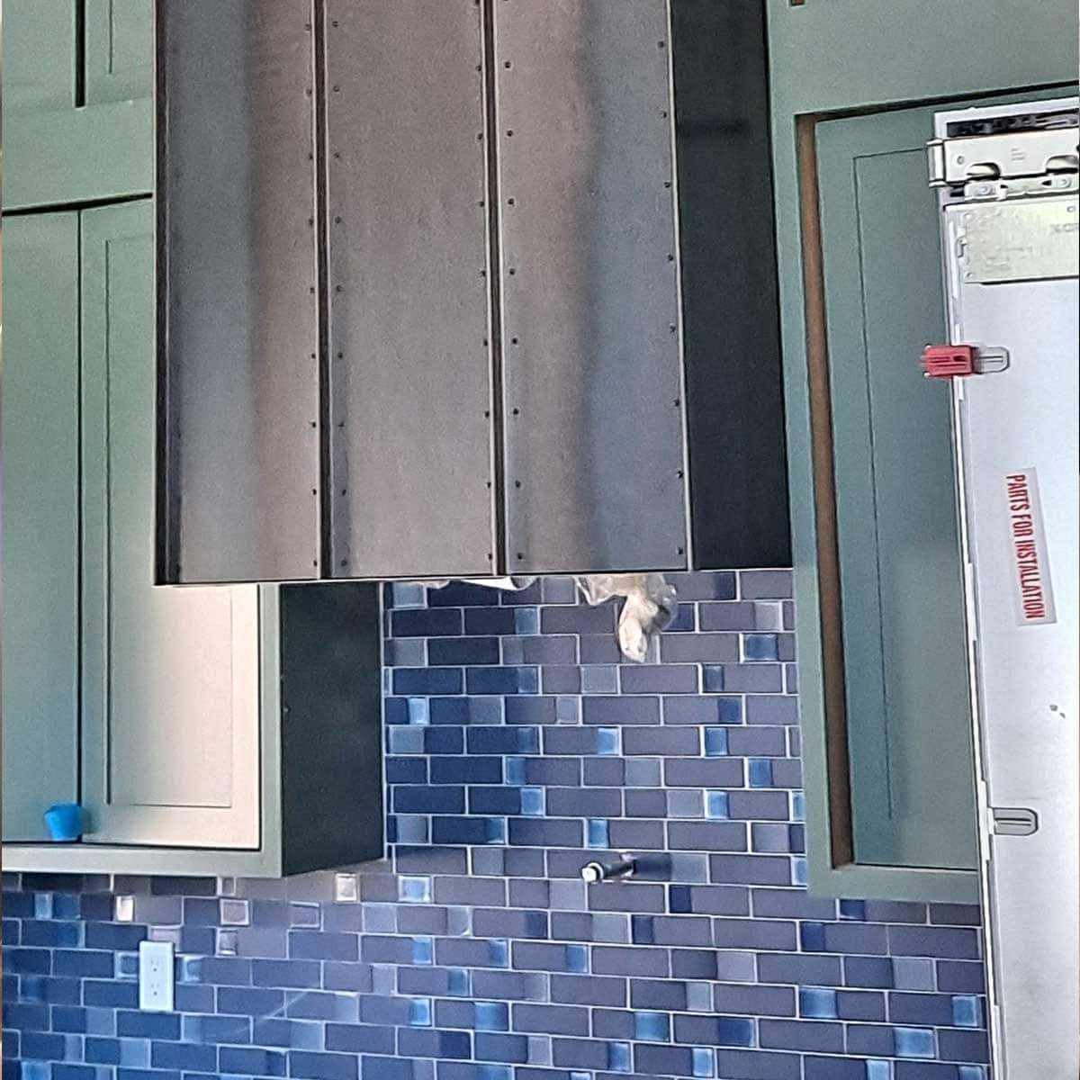 A kitchen with blue tile backsplash, green cabinets, a metal range hood, and a stainless steel appliance labeled "proof and holding cabinet.