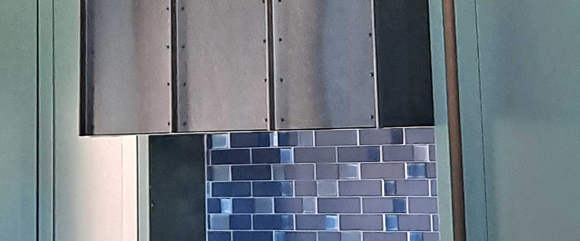 A section of a metallic vent or hood above a wall with blue and gray rectangular tiles.