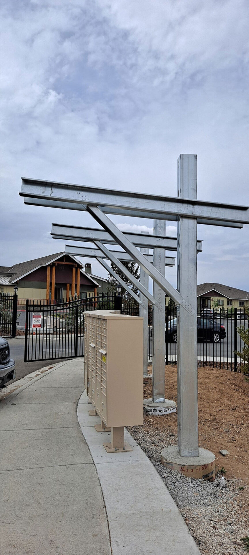 Rows of metal beams and mailbox units stand along a sidewalk next to a fenced residential area under a cloudy sky.