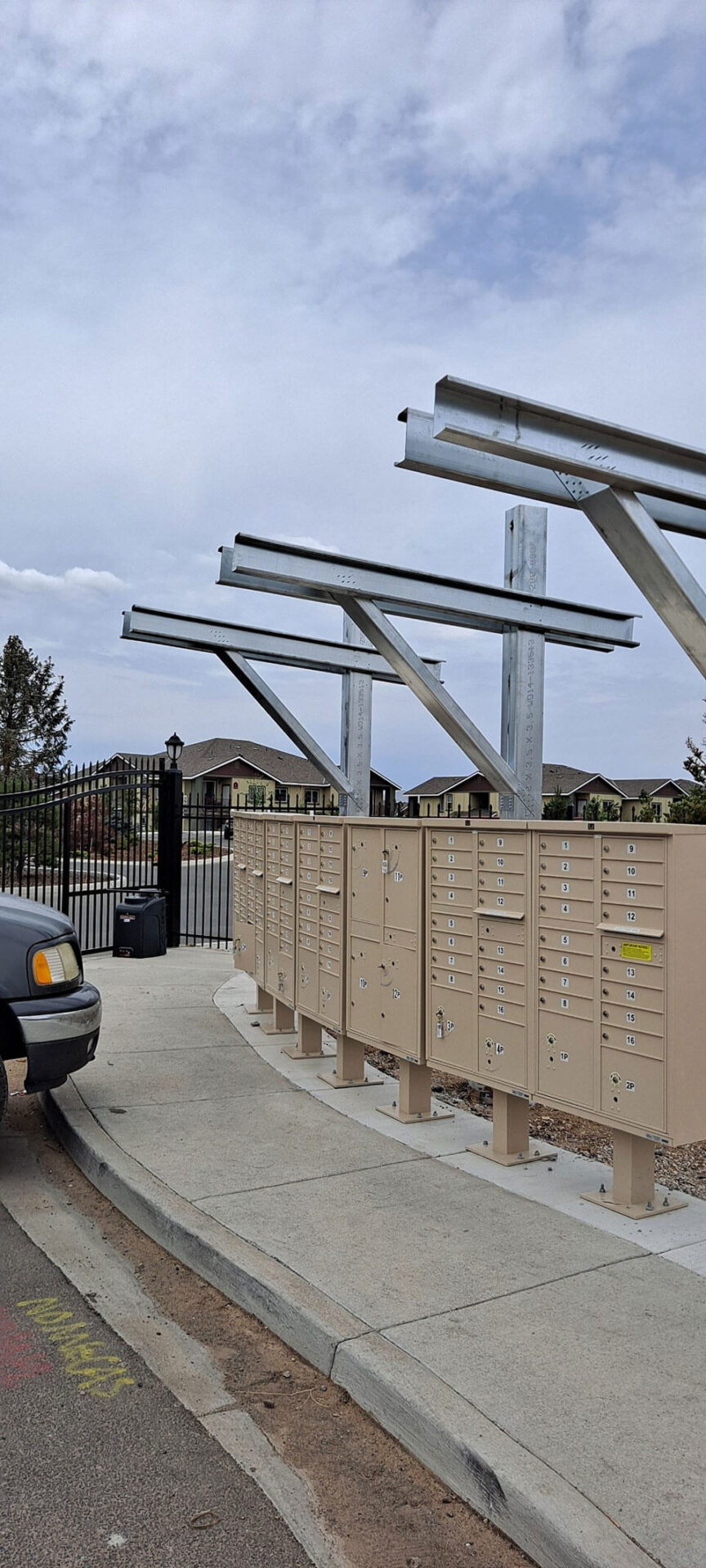 Tan cluster mailboxes are installed along a sidewalk, with unfinished metal frames overhead and a car parked nearby. A gated residential area is visible in the background.