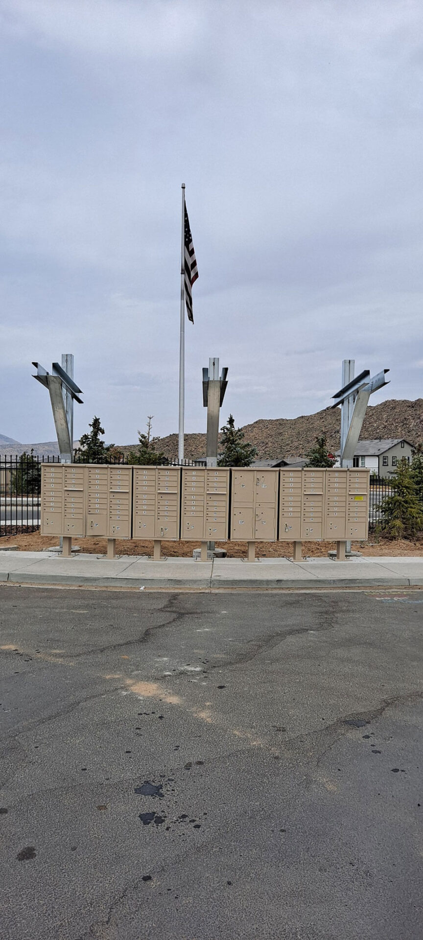 A row of beige community mailboxes is situated in front of a fence, with a U.S. flag on a pole and two modern metal sculptures in the background.
