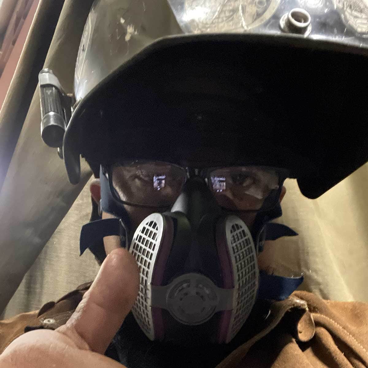 Person wearing a welding helmet, safety glasses, and a respirator mask gives a thumbs up.