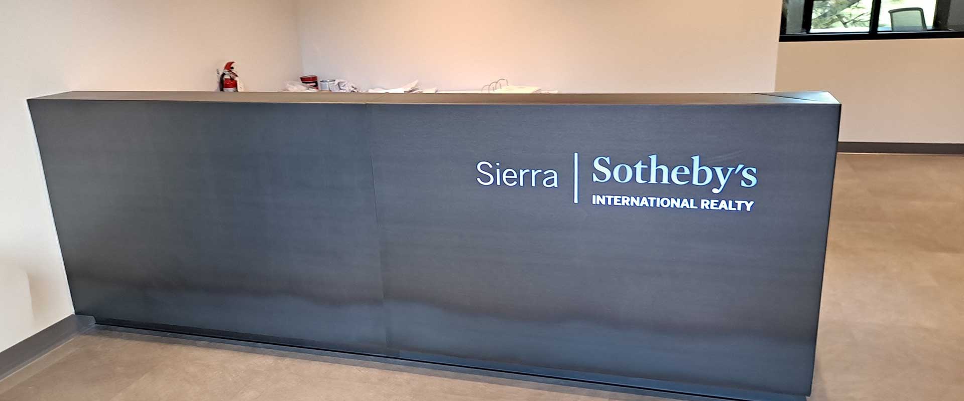Reception desk with "Sierra Sotheby's International Realty" sign on the front, located in a minimalistic office interior with beige walls and a fire extinguisher in the background.