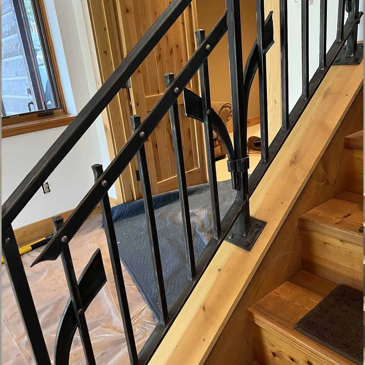 A wooden staircase with a black metal railing featuring decorative geometric shapes, next to a door and window, with protective coverings on the floor.