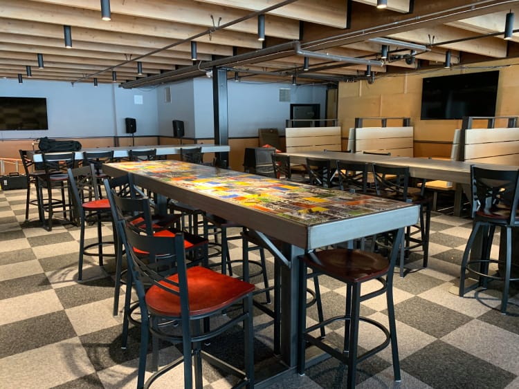 A modern, industrial-style restaurant interior with a long, colorful mosaic table, surrounded by black chairs with red seats, amidst a checkered black and grey floor.