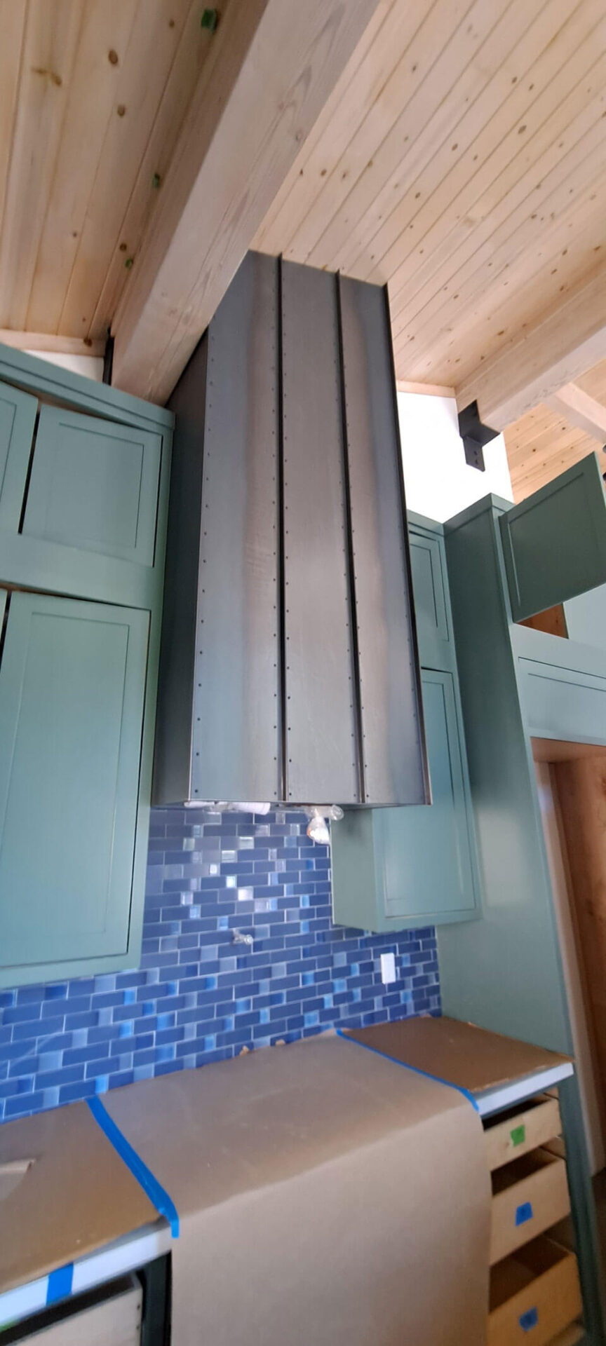 A metallic kitchen range hood installed above green cabinets and a blue tile backsplash, with a wood-paneled ceiling overhead.