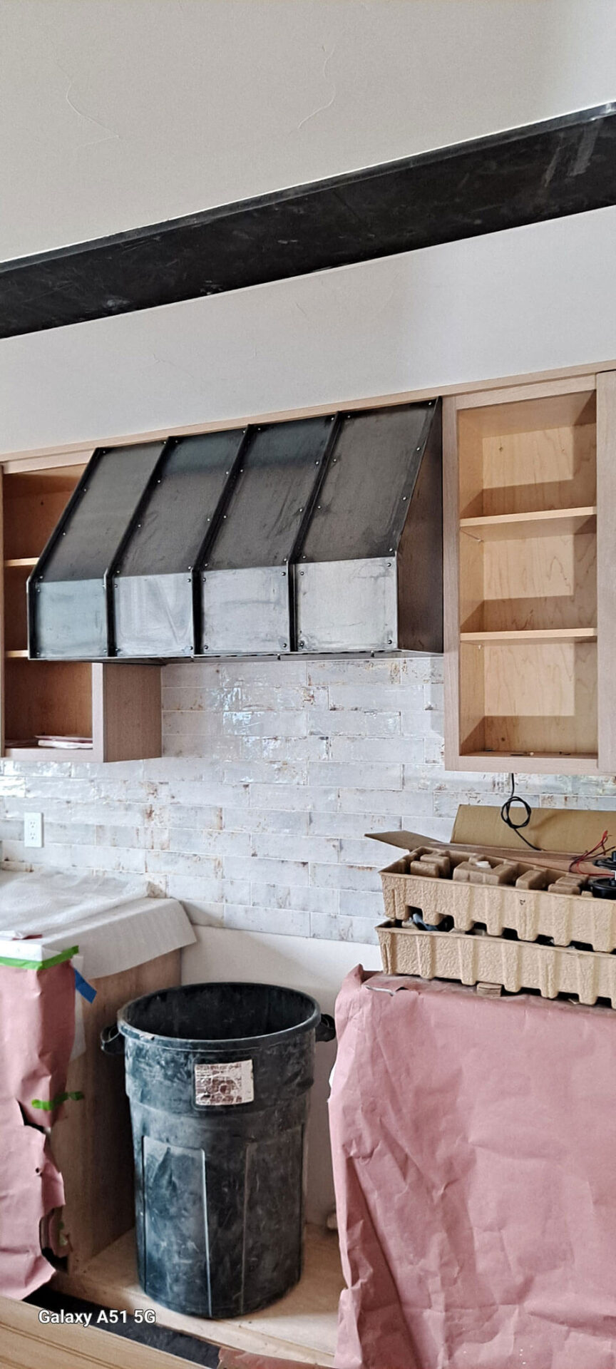 A kitchen under construction with unfinished wooden cabinets, a metal range hood, a trash bin, and surfaces covered with protective paper.