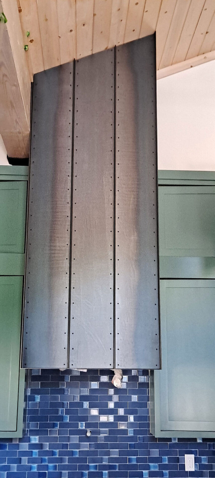 Large metal range hood with rivet details is installed above a blue tile backsplash and between green kitchen cabinets under a wooden ceiling.