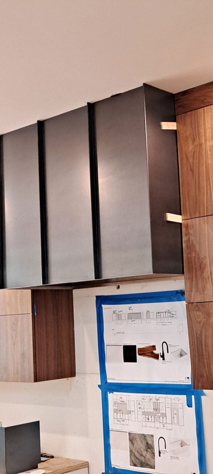 Metal and wood kitchen cabinets are installed above a countertop, with design plans taped to the wall below.