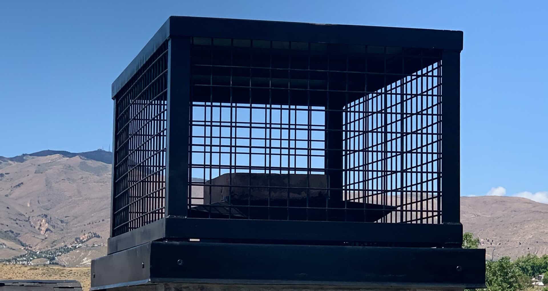 A black metal cage sits outdoors atop a platform with a mountainous landscape in the background under a clear, blue sky.