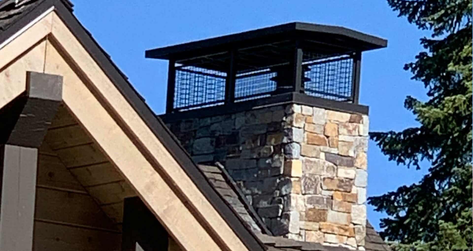 A stone chimney with a black metal cap and mesh sides is situated on a rooftop against a clear blue sky and a partial view of a tree.