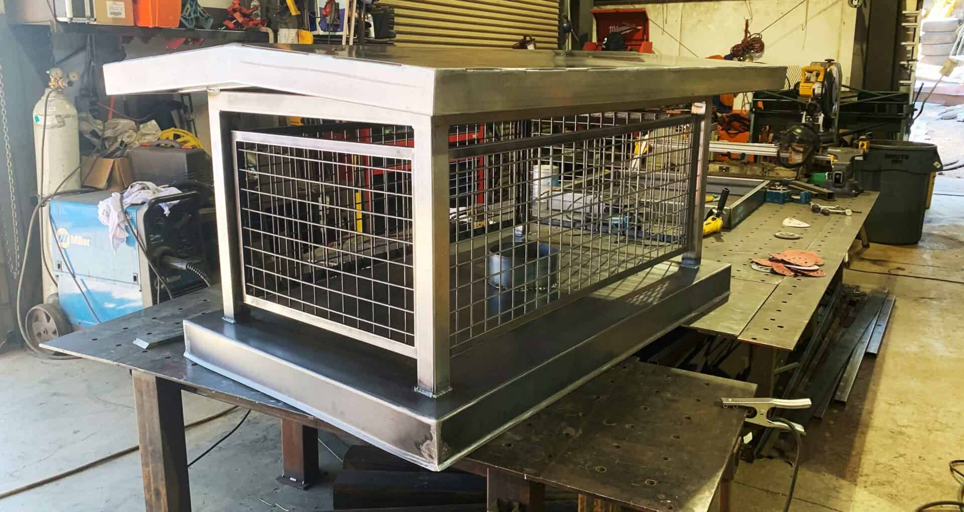 A large, rectangular metal structure with wire mesh sides sits on a cluttered workbench in a workshop. Tools, equipment, and materials are scattered around the workstation.