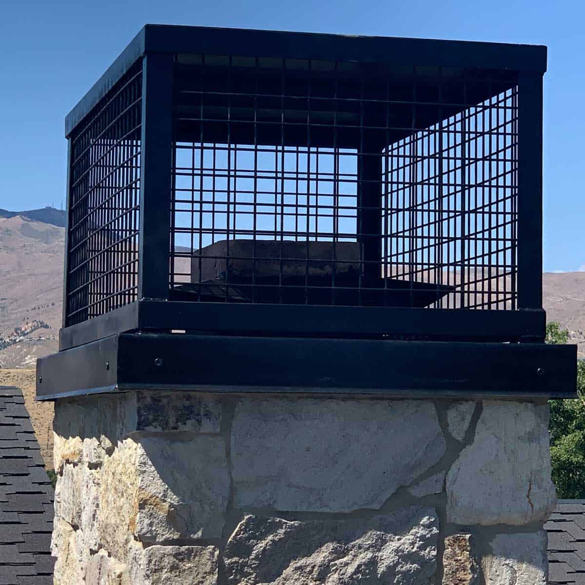 A stone chimney topped with a metal mesh cage, likely serving as a spark arrestor, with a mountainous landscape in the background.