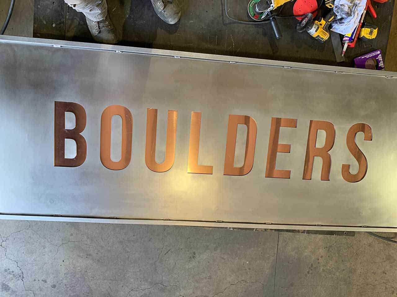 A metallic sign with the word "BOULDERS" inscribed in large copper letters is placed on a concrete floor. Tools and materials surround the sign.