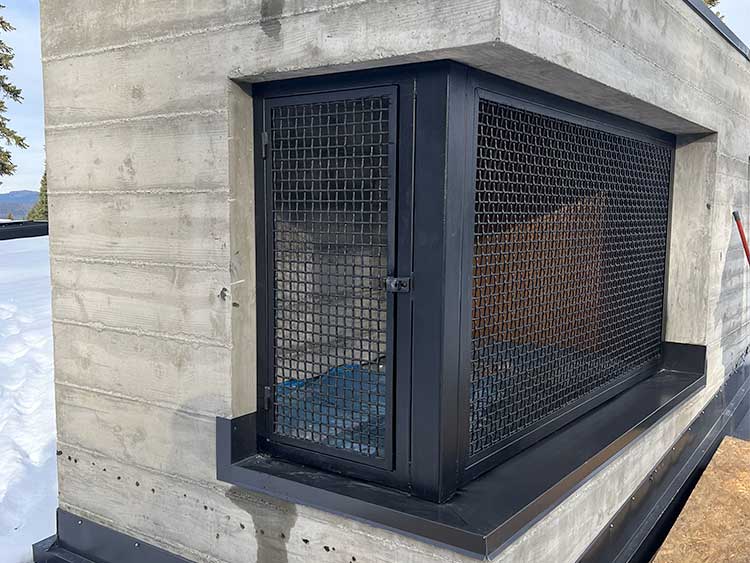 a concrete structure with a black metal mesh door and window, both secured and set into the wall, with snow visible on the ground nearby.