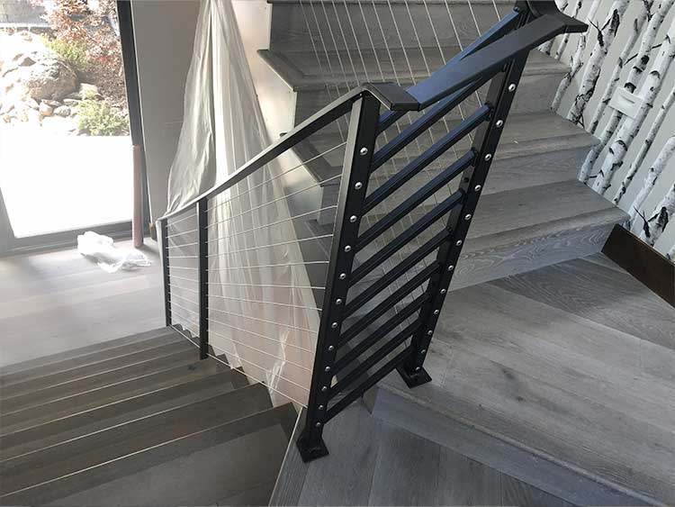 modern indoor staircase with light wood steps, metal cable railing, and black posts next to a wall with a birch tree pattern. natural light comes from a glass door on the left.