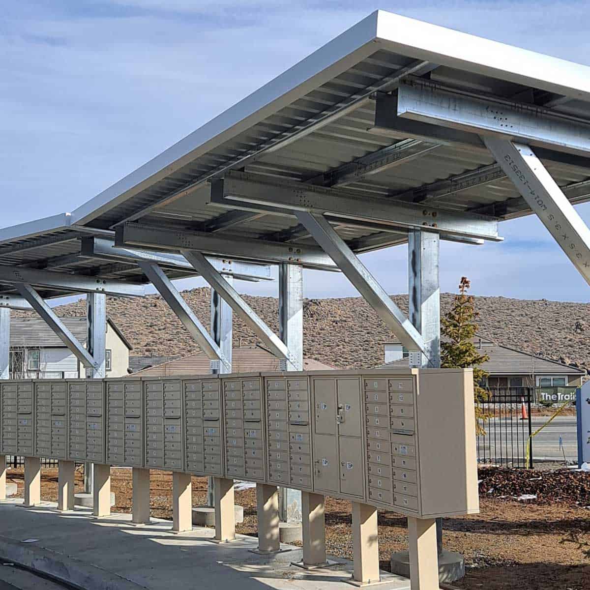 A row of outdoor mailboxes under a metal shelter with a sloping roof, situated in a residential area with a sign in the background.