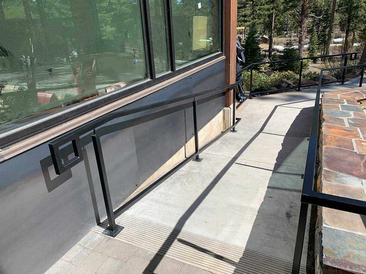 A concrete wheelchair ramp with a black handrail runs alongside a building with large windows and leads to a paved area.