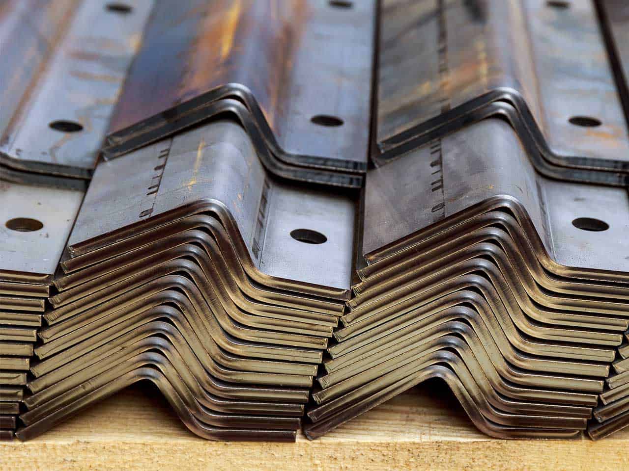 Neatly stacked metal brackets with a zigzag shape, featuring circular holes and slight rust patches, arranged on a wooden surface.