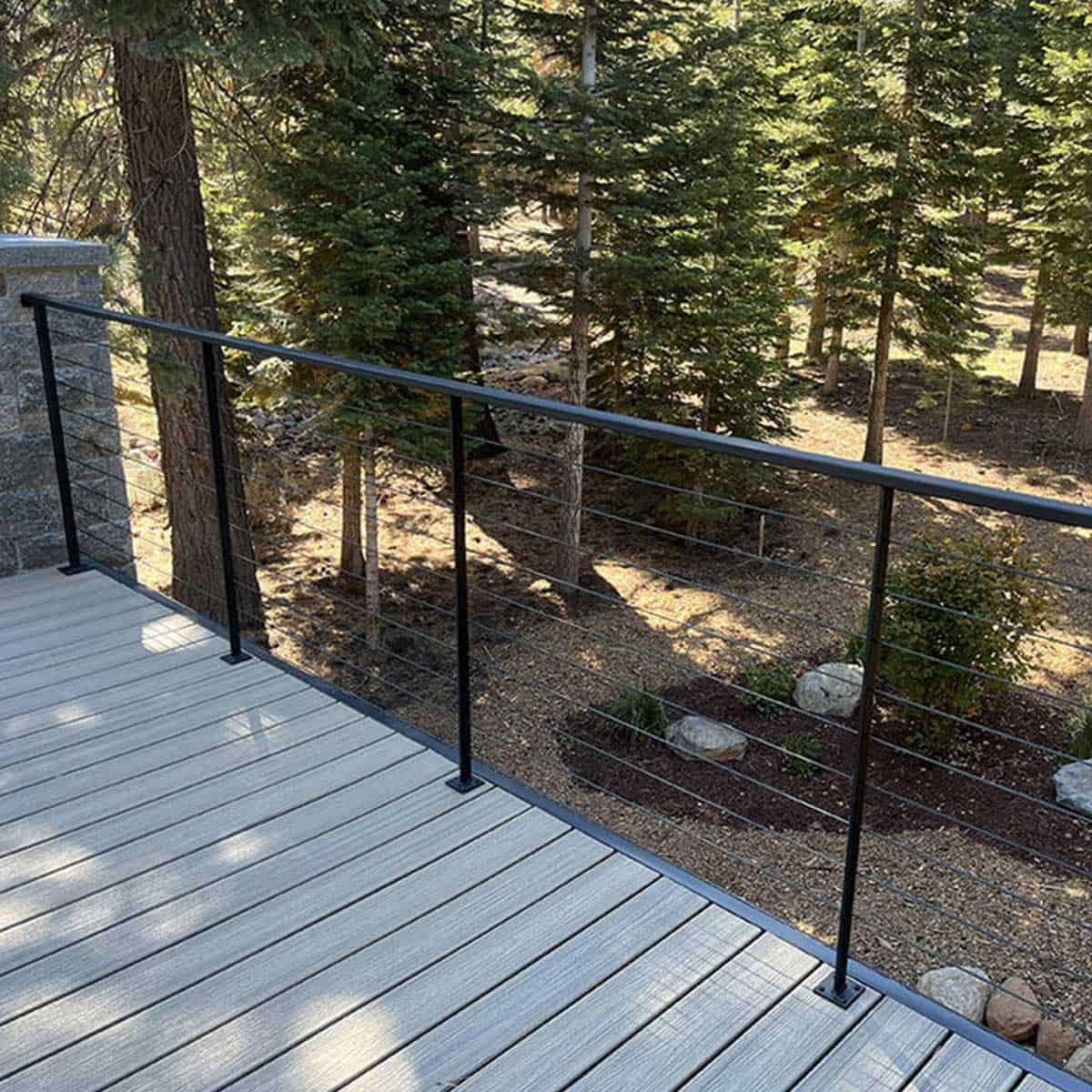 A deck with light-colored wooden flooring and a black metal railing overlooks a forested area with evergreen trees and scattered rocks.