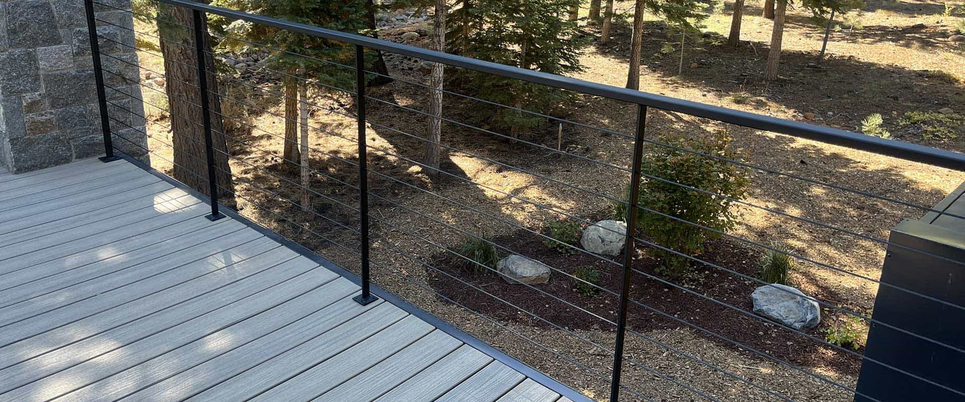 A deck with light-colored wood planks and a black metal cable railing overlooks a landscaped yard with rocks, mulch, and trees.