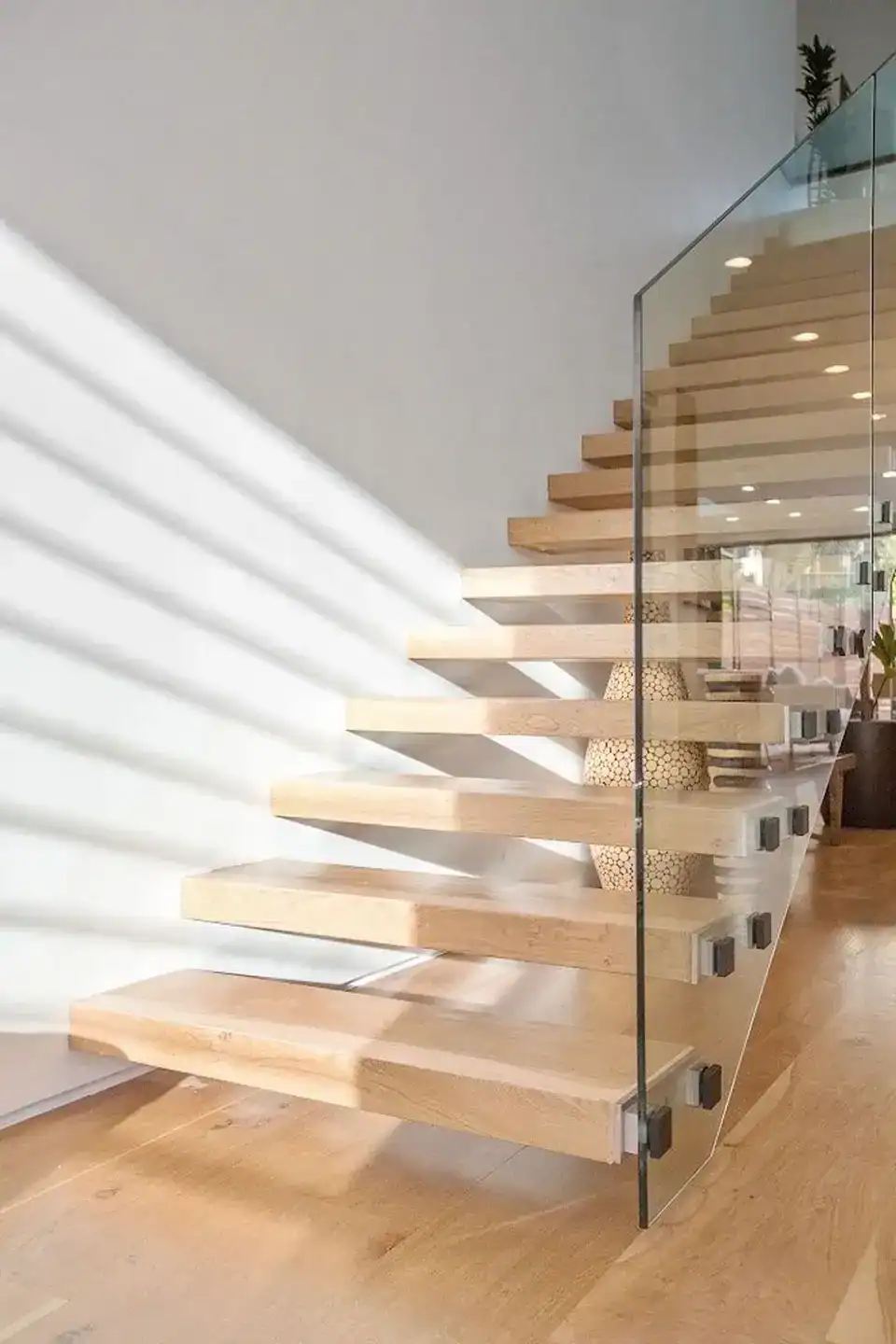 a modern floating wooden staircase with metal brackets and a glass railing, set against a white wall with sunlight casting shadows.