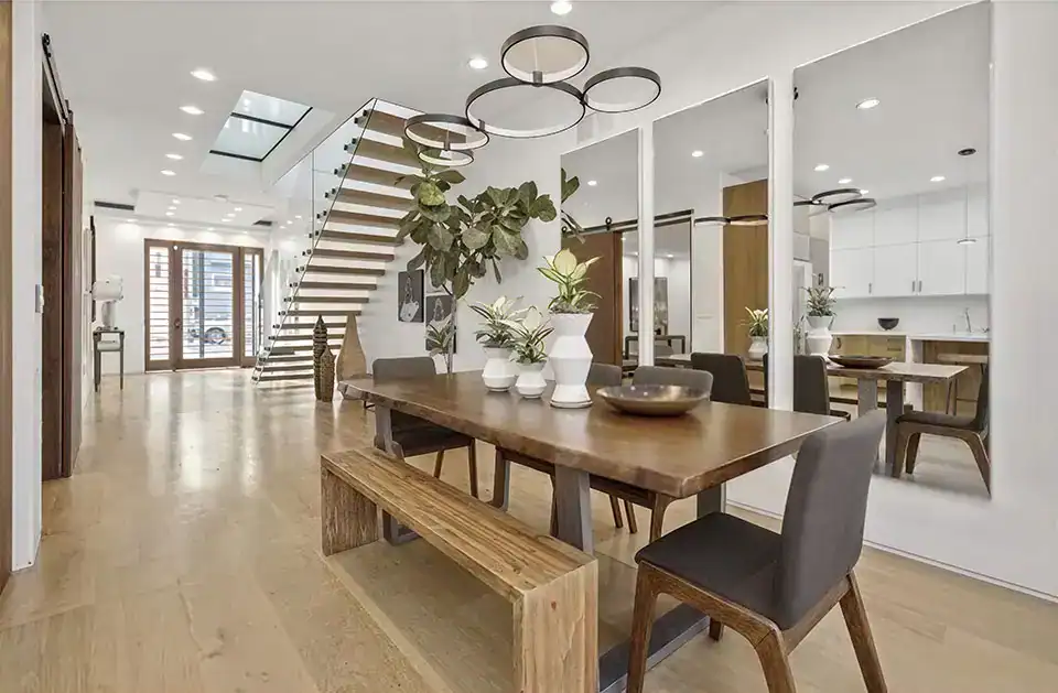 modern dining area with wooden table, bench seating, black chairs, potted plants, large mirrors, floating staircase, and open view to a bright kitchen.