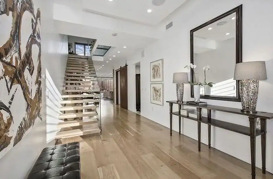 modern hallway with wooden floors, floating staircase, console table with two lamps, a large mirror, abstract wall art, and a black bench.