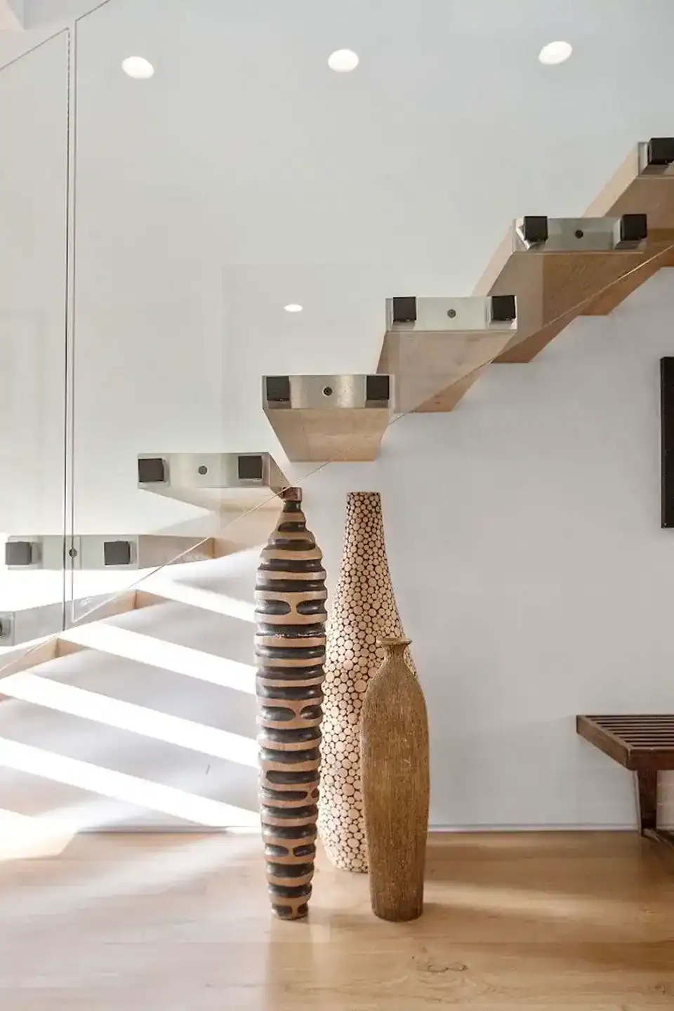 floating wooden stairs with glass railing, positioned above three decorative tall vases on a wooden floor, with sunlight casting shadows on the wall.